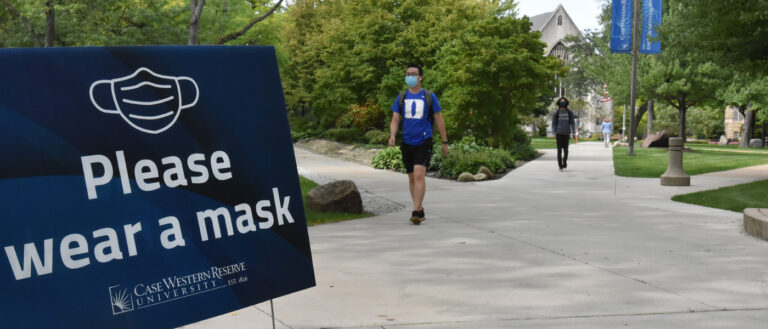 Photo of a sign that says "please wear a mask" next to a path on CWRU's campus with students wearing masks walking nearby