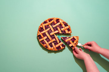 Photo from above of a person cutting a piece out of a berry pie against a plain background