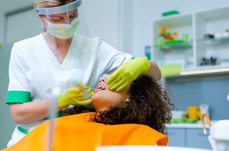 Mixed-race teenage girl with beautiful curly hair on dental checkup by middle-age caucasian woman. Coronavirus safety precaution at dentist's office.