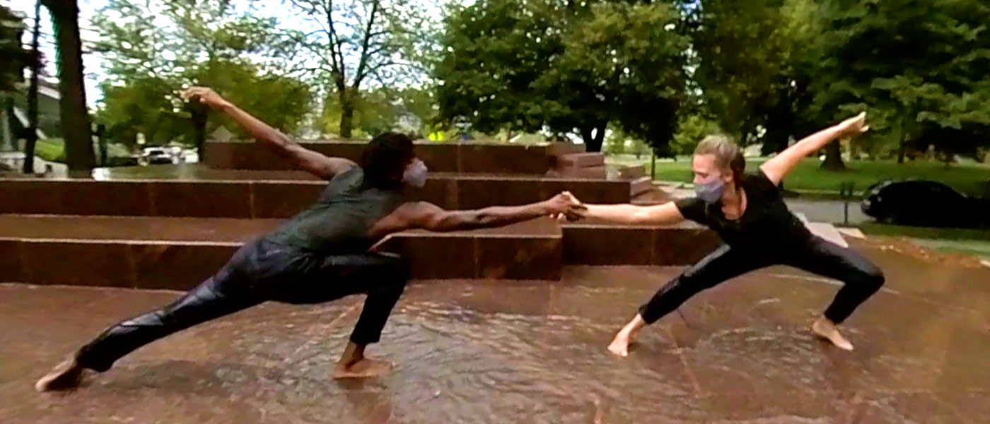 Photo of two dancers with outstretched hands holding each other on the merging fountain on campus