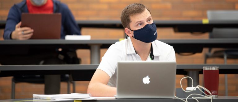 Photo of a masked student sitting a classroom with a laptop on the desk while looking up ahead