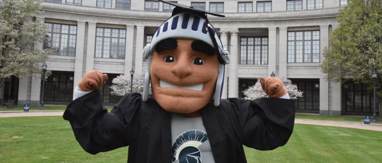 Photo of Spartie wearing a graduation cap and gown posing for a photo in front of Kelvin Smith Library