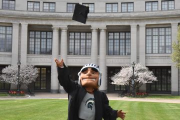 Photo of Spartie mascot wearing a graduation gown and tossing a cap in the air in front of Kelvin Smith Library