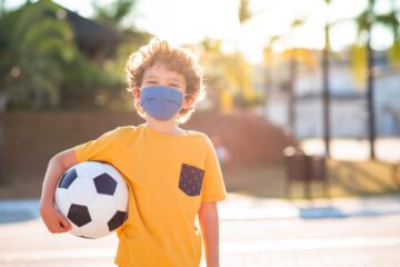 Child with facial tissue protection mask with soccer ball outdoors during pandemic