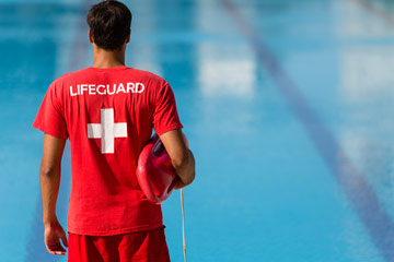 Photo of a lifeguard looking at a pool from behind wearing all read and a T-shirt that reads "lifeguard" and holding a floatation device