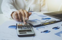 Close up of businessman or accountant hand holding pen working on calculator to calculate the number, graph chart, and business data. Accountancy documents and business information on the desk at the office, Business concept.