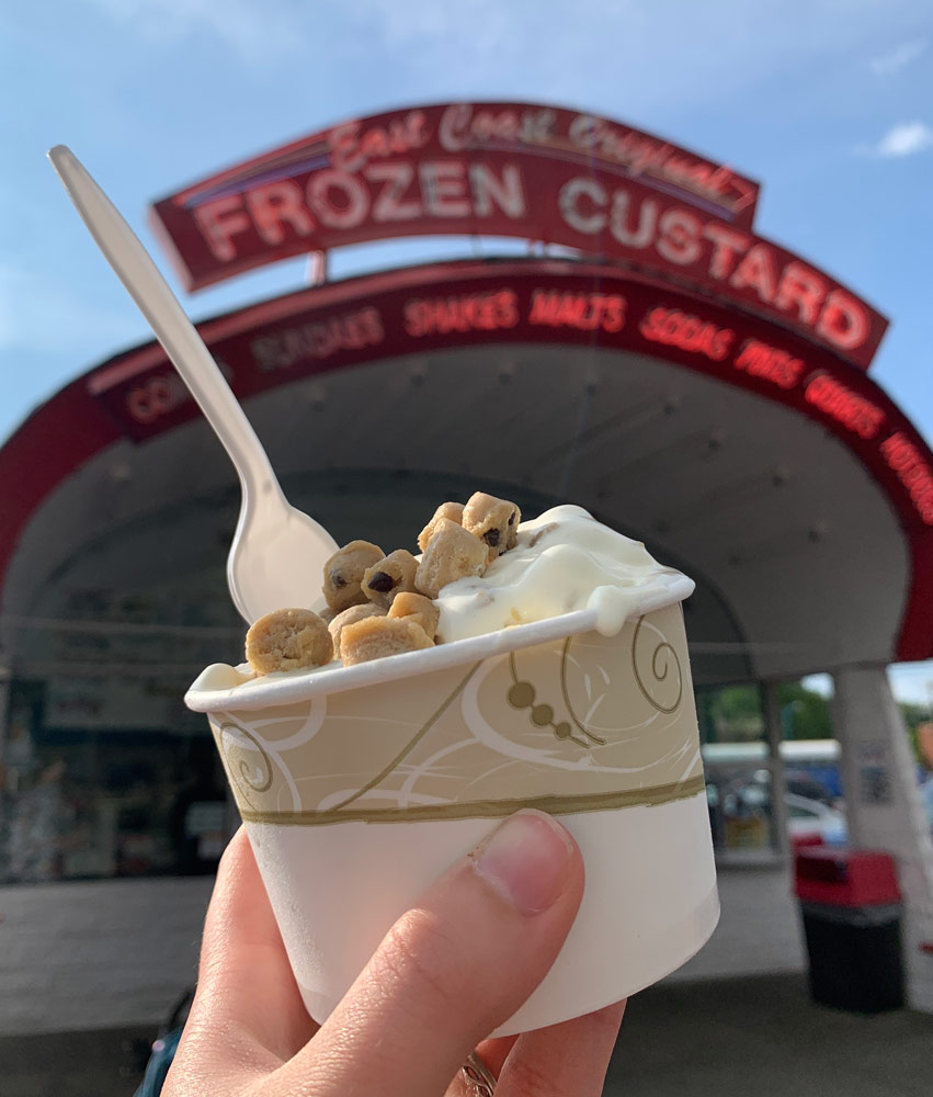 Photo of a hand holding up a cup of a cookie dough concrete in front of the East Coast Original Frozen Custard building's marquee-like sign