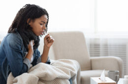 Sick Black Woman Coughing Hard At Home, Sitting On Couch Wearing Scarf And Covered With Blanket, Side View With Copy Space