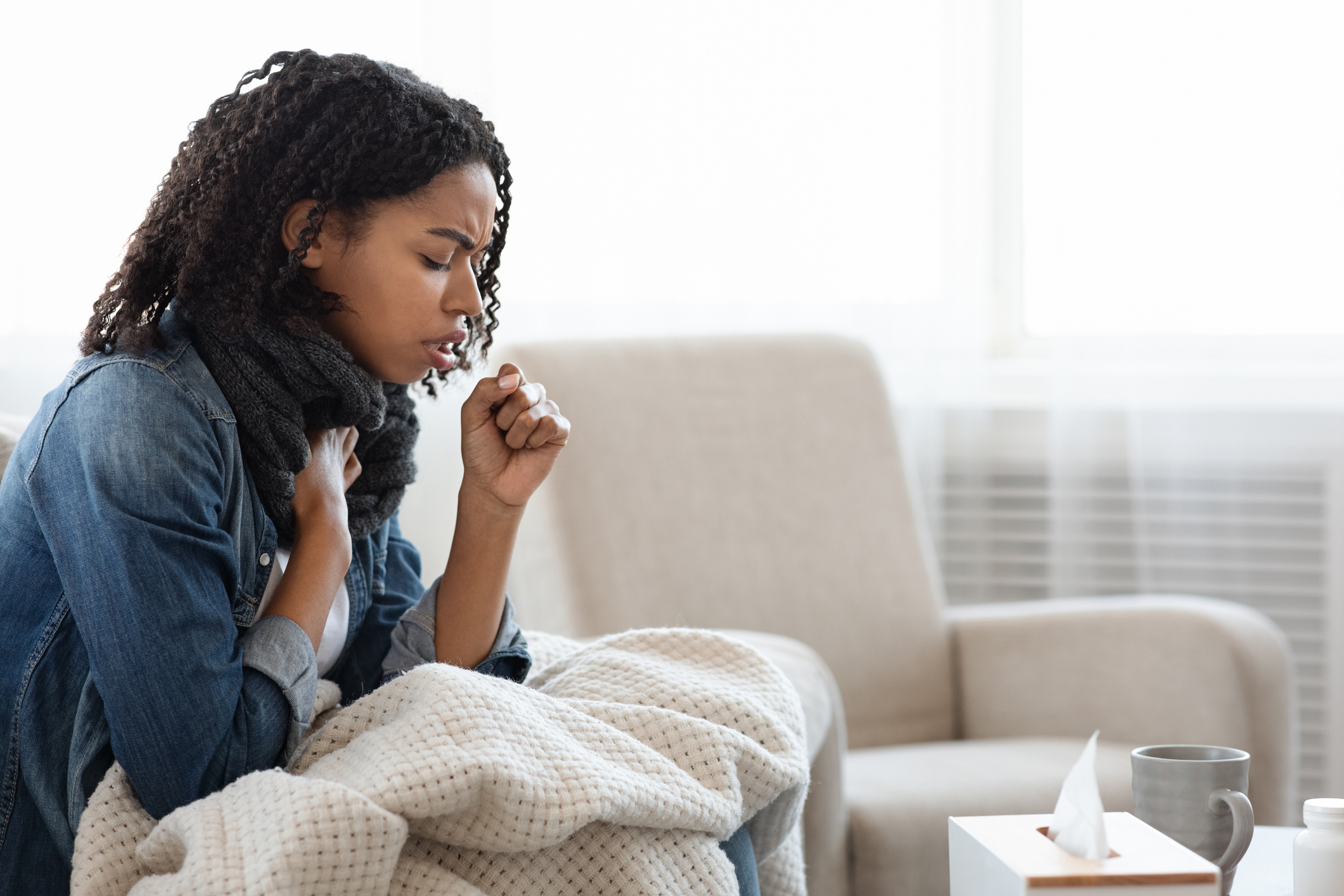 Sick Black Woman Coughing Hard At Home, Sitting On Couch Wearing Scarf And Covered With Blanket, Side View With Copy Space