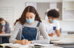 High school student taking notes from book while wearing face mask due to coronavirus emergency. Young woman sitting in class with their classmates and wearing surgical mask due to Covid-19 pandemic. Focused girl studying in classroom completing assignment during corona virus.