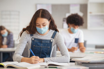 High school student taking notes from book while wearing face mask due to coronavirus emergency. Young woman sitting in class with their classmates and wearing surgical mask due to Covid-19 pandemic. Focused girl studying in classroom completing assignment during corona virus.