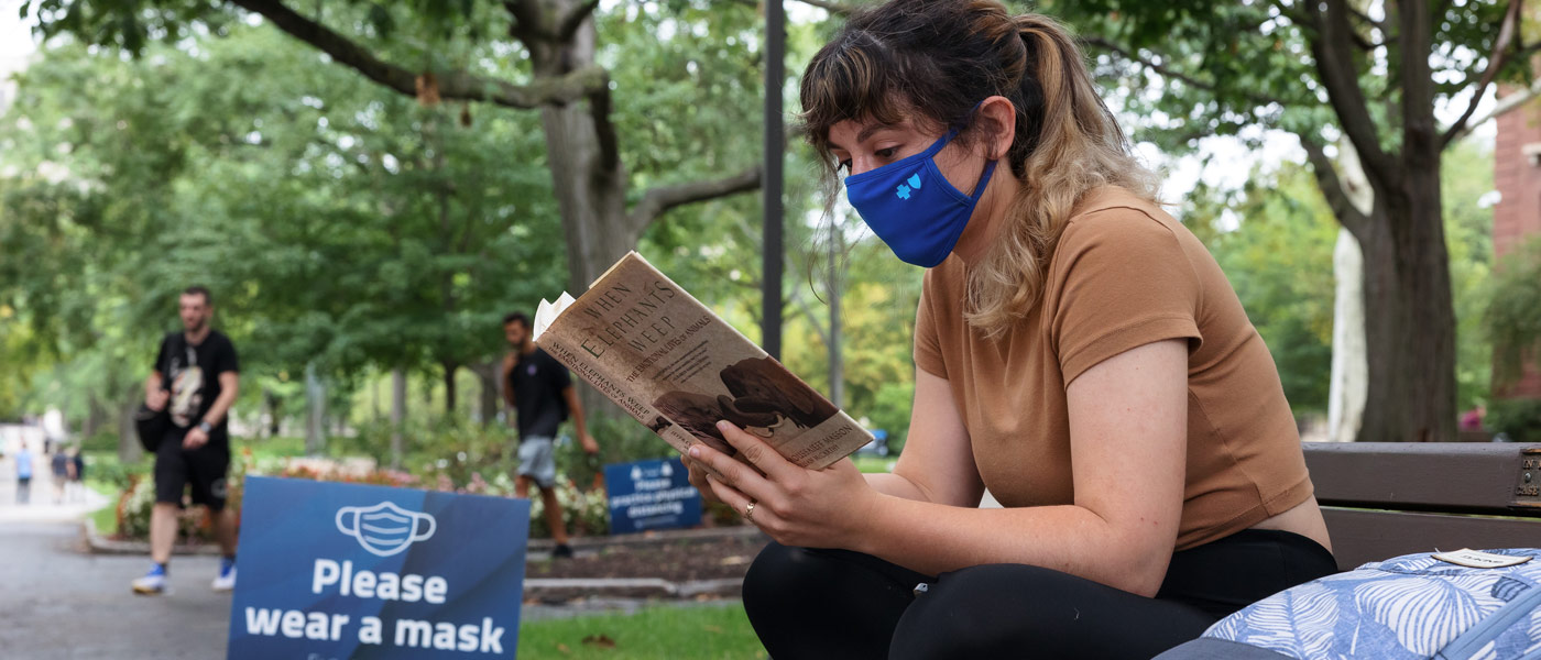 Photo of a masked student reading a book on a bench next to a sign that says "please wear a mask"
