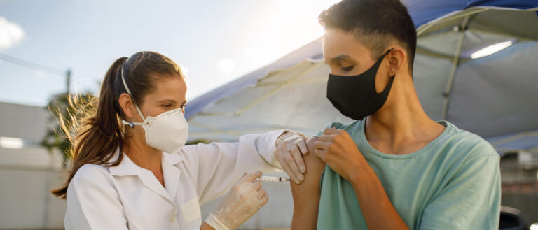 Photo of a health care professional administering a vaccine to a teenage boy while both wear masks