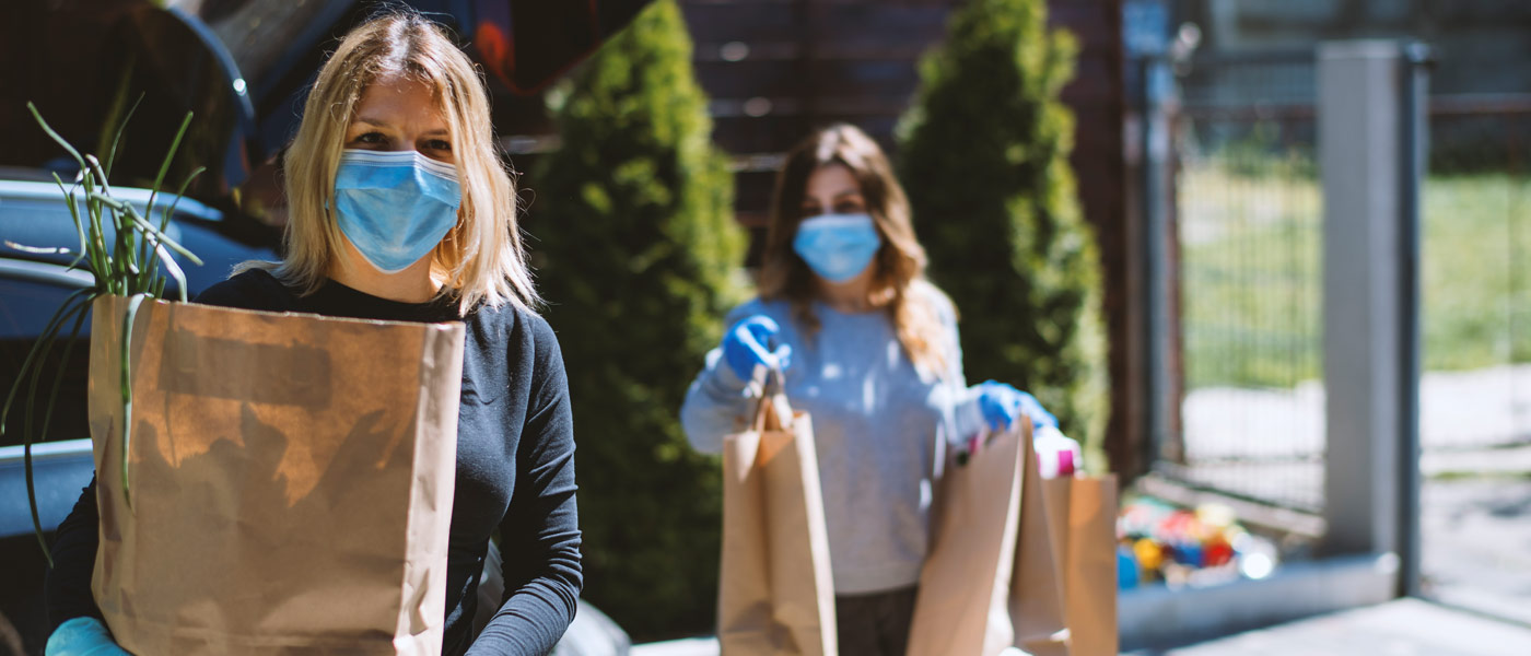 Photo of two women wearing masks and gloves holding paper bags of groceries