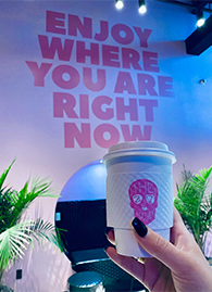 A hand holding a 27 Club Coffee to-go cup in front of a wall decal that reads "enjoy where you are right now" 