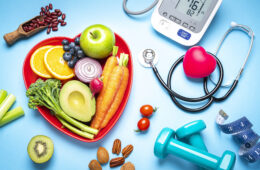 Red heart shape plate with fresh organic fruits and vegetables shot on blue background. A digital blood pressure monitor, doctor stethoscope, dumbbells and tape measure are beside the plate