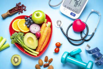 Red heart shape plate with fresh organic fruits and vegetables shot on blue background. A digital blood pressure monitor, doctor stethoscope, dumbbells and tape measure are beside the plate