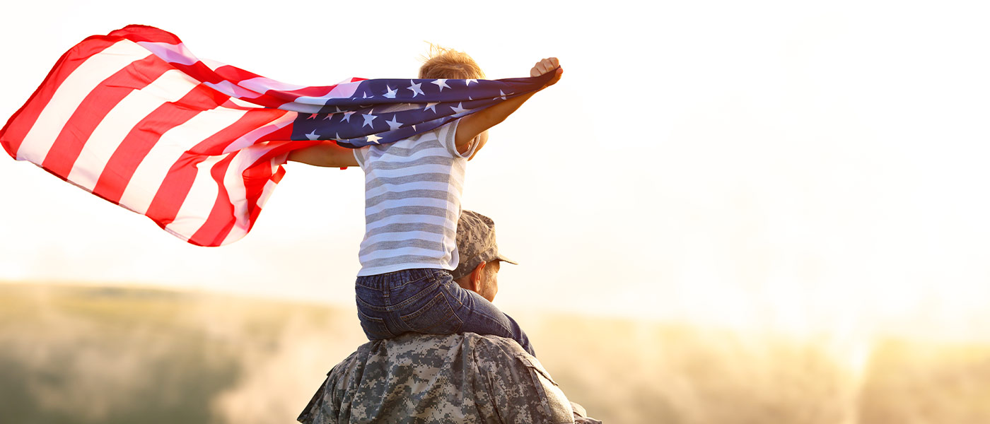 Photo of a child on a veteran's shoulders holding an American flag like a cape