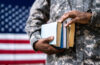 Photo of someone wearing Army fatigues and holding books with an American flag in the background