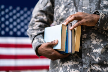 Photo of someone wearing Army fatigues and holding books with an American flag in the background