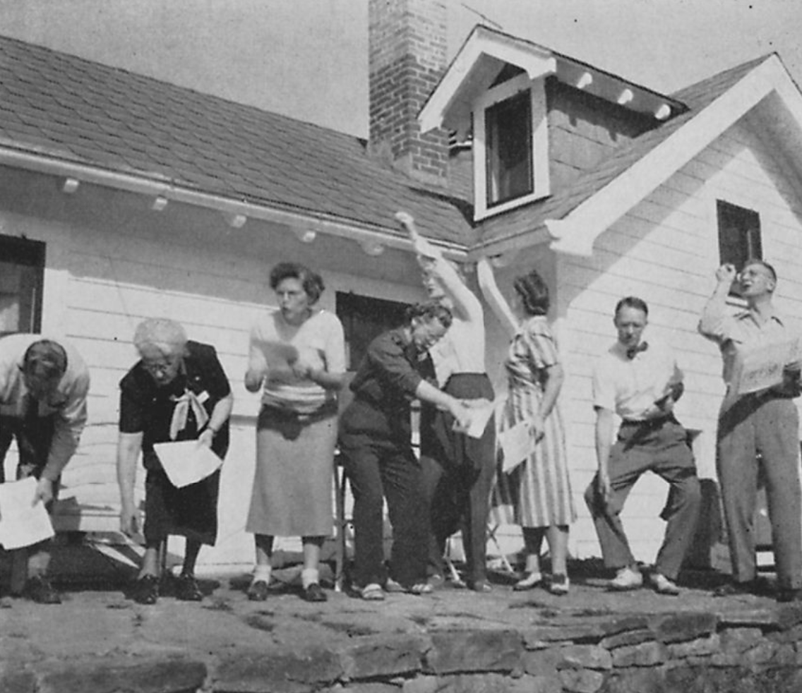 black and white view of 8 people wearing casual attire, standing in various poses while holding pieces of paper / building in background / retaining wall in foreground