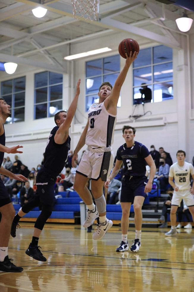 Photo of men's basketball player Mitch Prendergast holding a basketball in his outstretched hand to go for a shot while an opposing player attempts to block it