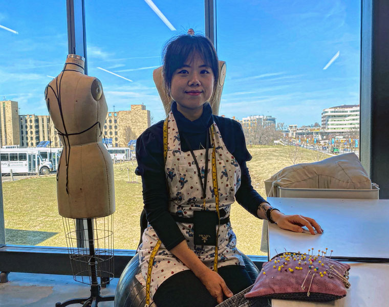 Photo of CWRU costume shop manager Rainie Jiang sitting in the costume shop in front of a dress form and floor-to-ceiling windows