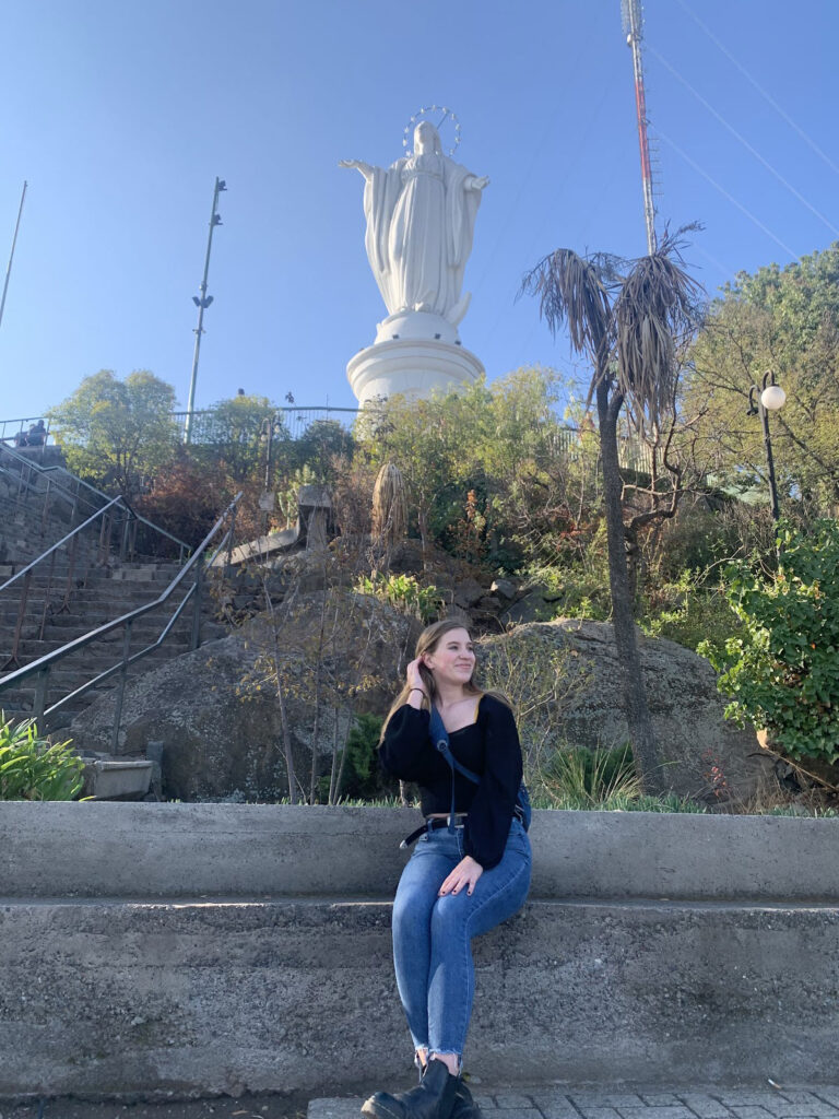 Photo of Delphine Clatanoff sitting along a wall at the top of Santiago’s Cerro (Hill) San Cristóbal with the virgin statue above