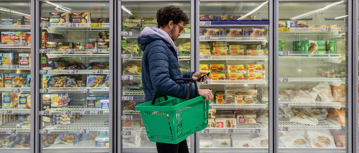 Photo of a man checking his phone and a shopping basket while standing next to a case of food in a grocery store