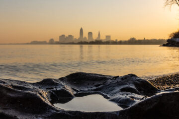 Photo of Lake Erie during a sunrise with a rocky coastline in the foreground and the Cleveland skyline in the background