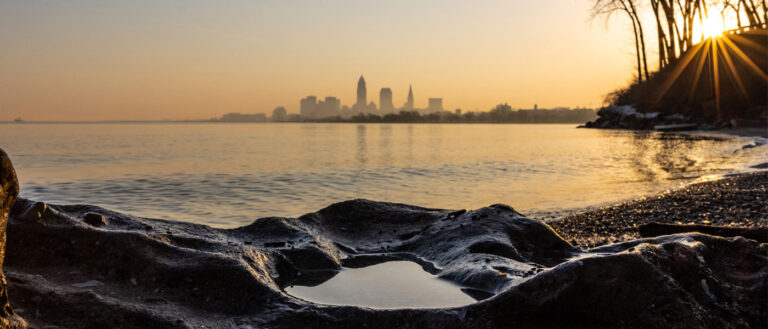 Photo of Lake Erie during a sunrise with a rocky coastline in the foreground and the Cleveland skyline in the background