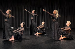 Photo of six dancers all dressed in black with three standing with an arm outstretch and the other three sitting on the ground and holding onto the legs of the others