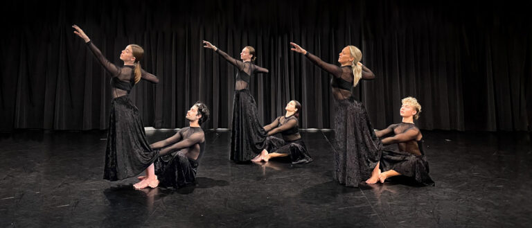 Photo of six dancers all dressed in black with three standing with an arm outstretch and the other three sitting on the ground and holding onto the legs of the others