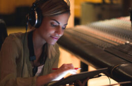 Photo of a woman with headphones on working on a tablet in a studio and a music mixing board behind her