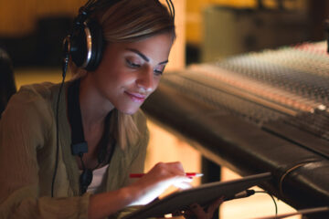 Photo of a woman with headphones on working on a tablet in a studio and a music mixing board behind her