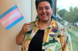 Photo of Harley French happily waving the transgender pride flag while wearing bisexual and non-binary pride flag pins.