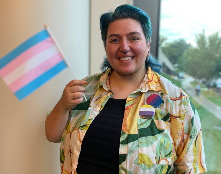 Photo of Harley French happily waving the transgender pride flag while wearing bisexual and non-binary pride flag pins.