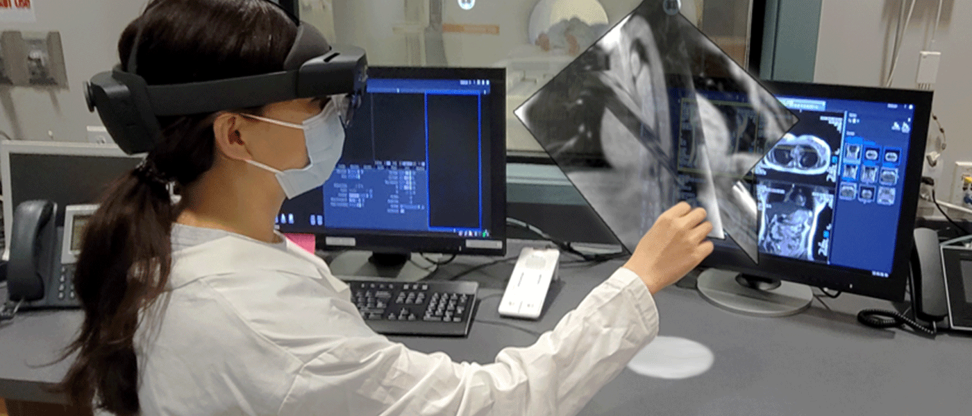 a woman wearing a mixed-reality headset viewing a computer screen showing a patient inside an MRI