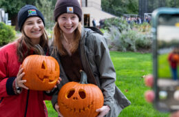 Photo taken from over someone's shoulder showing them taking a photo of two CWRU students holding carved pumpkins
