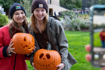 Photo taken from over someone's shoulder showing them taking a photo of two CWRU students holding carved pumpkins