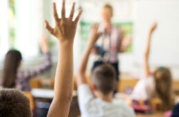 photo of students from behind in a classroom with one student prominently raising a hand