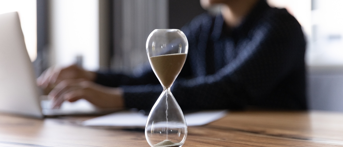 Close up of an hourglass on a wooden office table as a busy employee works on laptop in the background