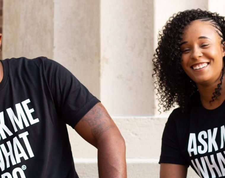 Two people smile while wearing black shirts and holding cards that say "Ask me what I do"