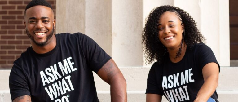 Two people smile while wearing black shirts and holding cards that say "Ask me what I do"