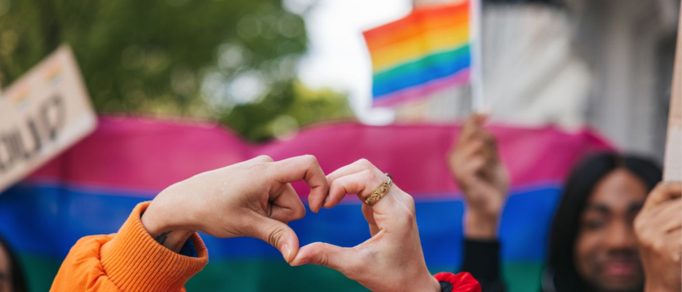 Genderqueer and non-binary friends making a heart shape against a rainbow flag celebrating love during a Pride Parade.