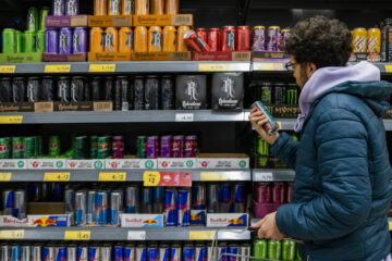Photo of a man looking at the back of a can of an energy drink in front of a case of caffeinated beverages