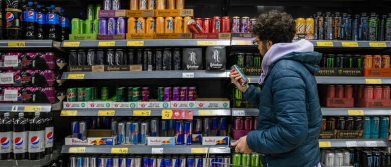 Photo of a man looking at the back of a can of an energy drink in front of a case of caffeinated beverages