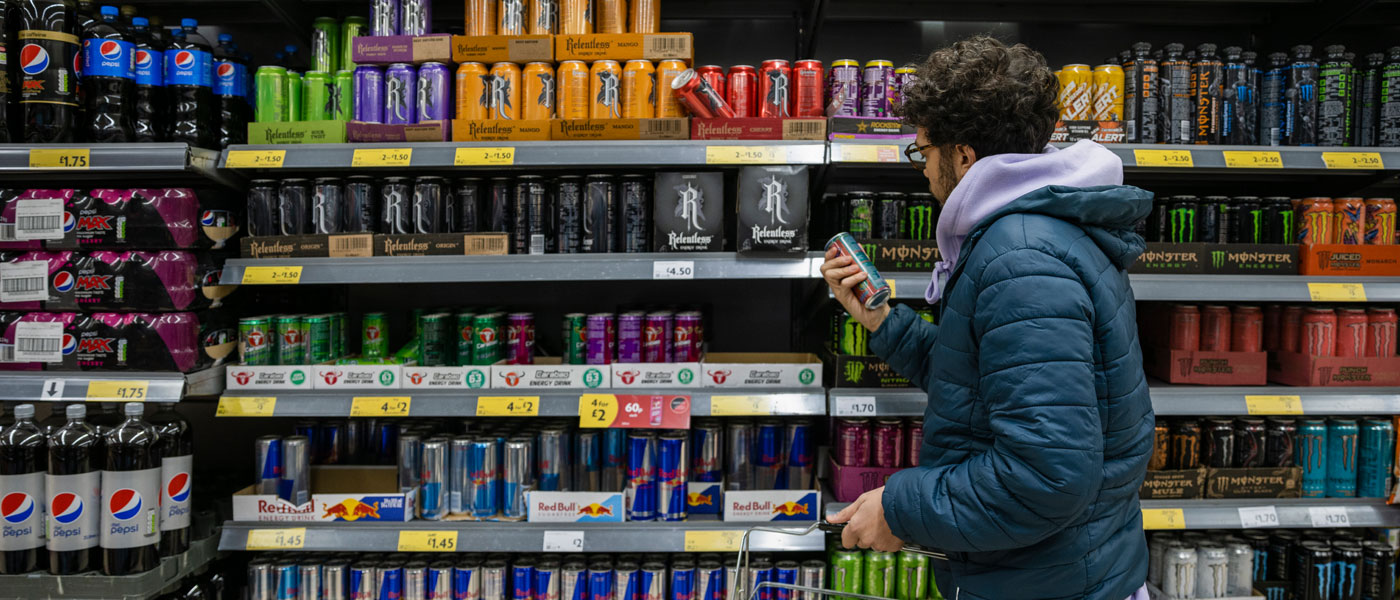 Photo of a man looking at the back of a can of an energy drink in front of a case of caffeinated beverages