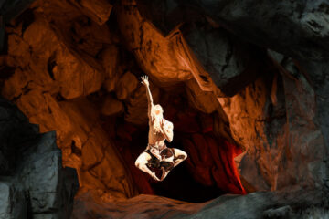 Photo of a dancer leaping on stage with an arm extended upwards with a cave-like backdrop behind them