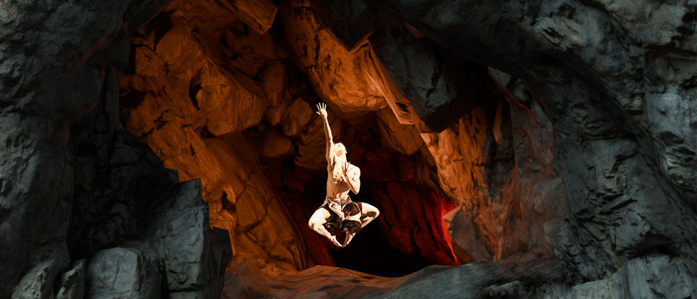 Photo of a dancer leaping on stage with an arm extended upwards with a cave-like backdrop behind them